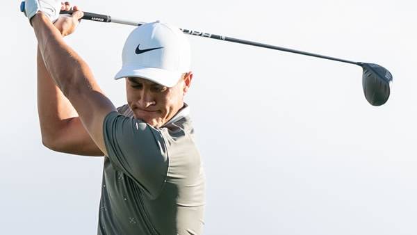Male golfer in a white Nike cap and grey shirt at the top of his golf swing.
