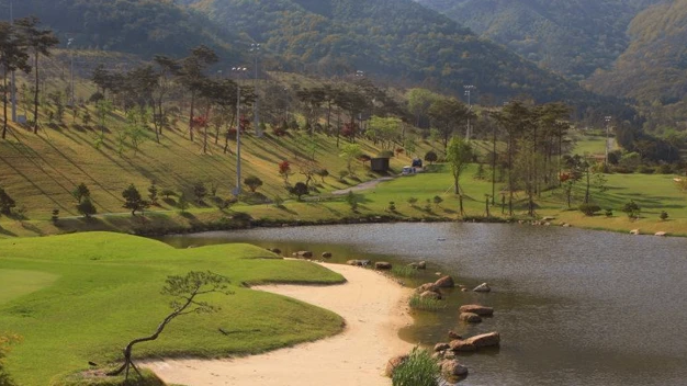 A golf course with a lake, sand trap, green hills, and tree-covered mountains.