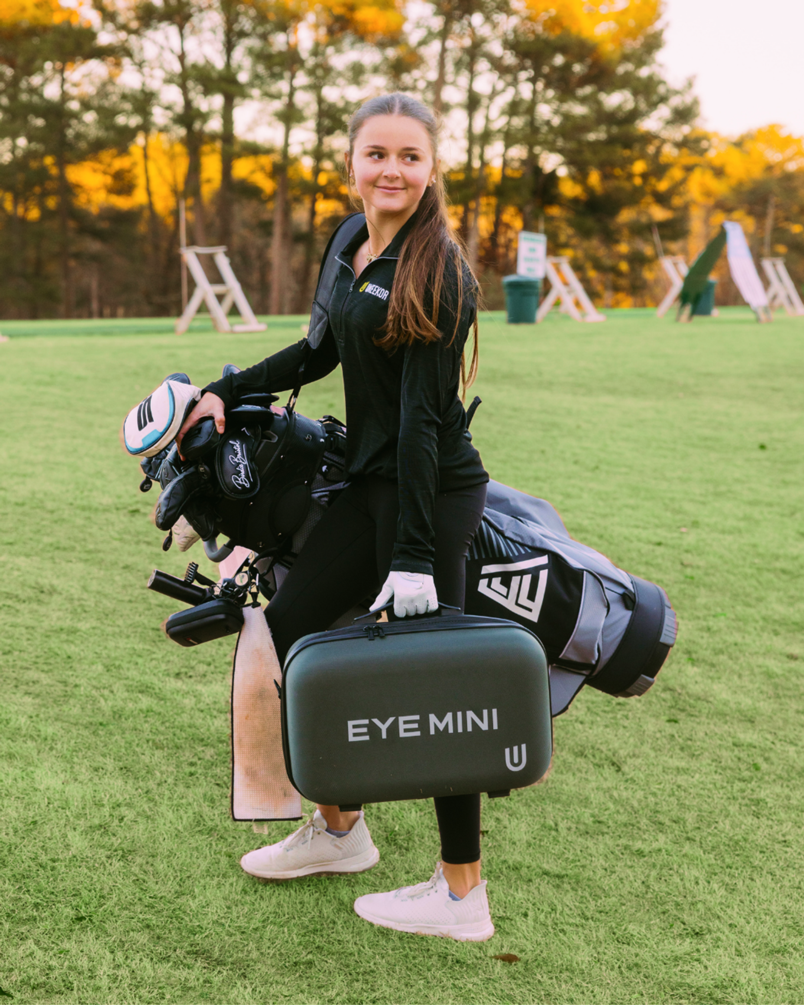 A young woman on a golf course carrying a golf bag and holding an "EYE MINI" case.
