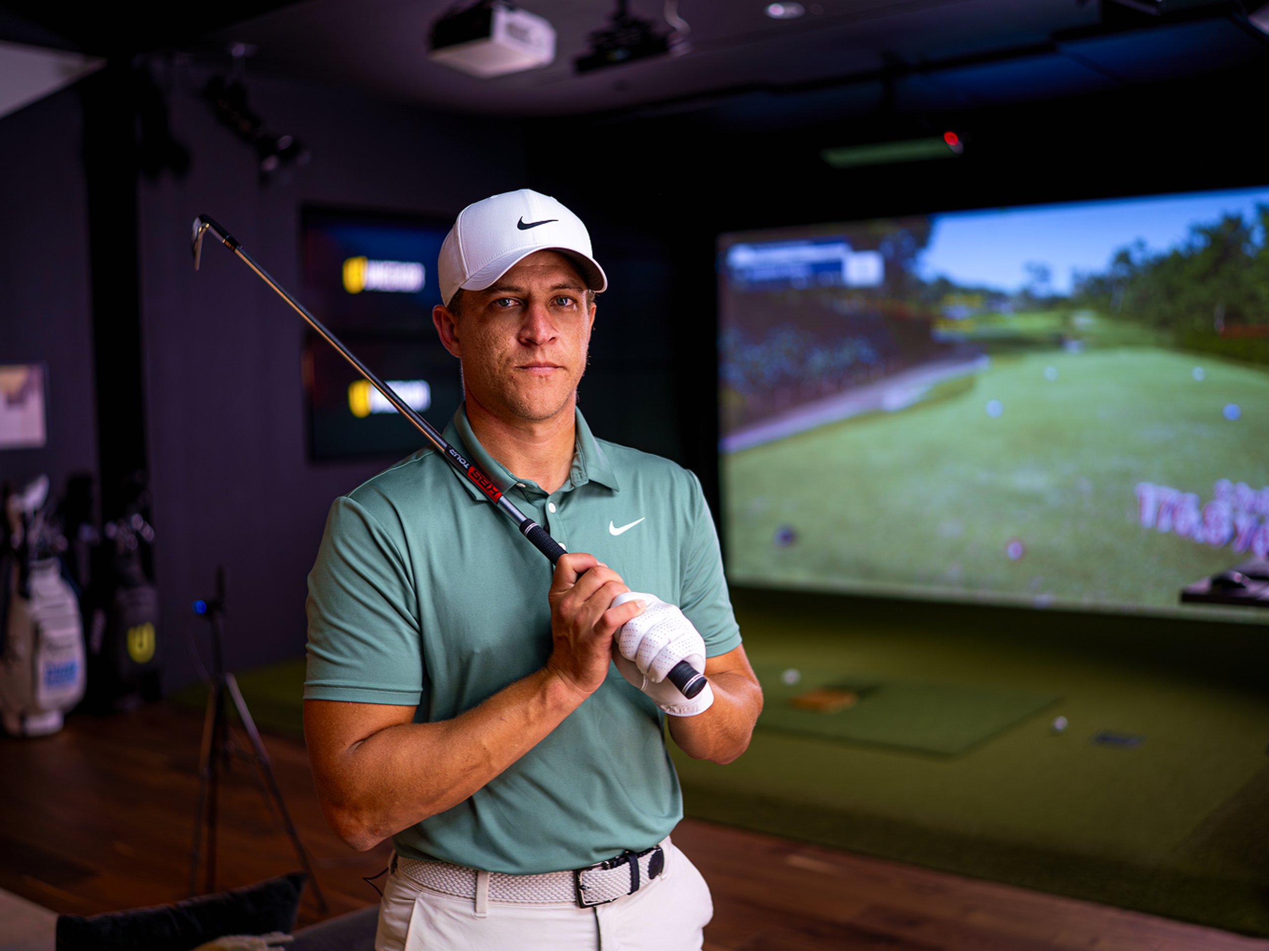 A man in golf attire holds a club and looks at the camera inside a golf simulator.