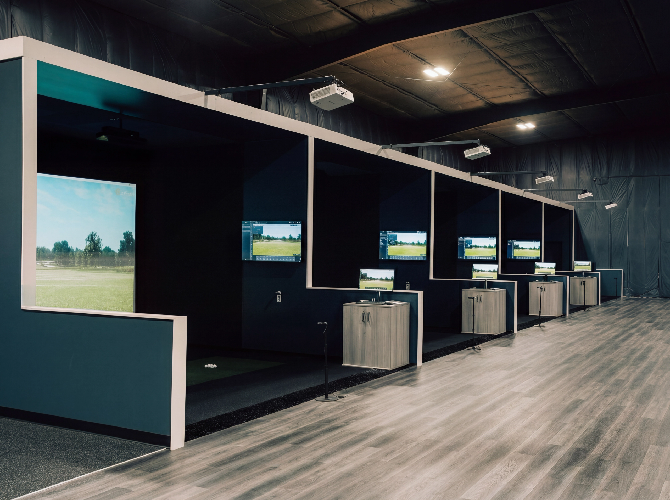 A row of indoor golf simulator bays, each with a large screen showing a golf course, a hitting mat, and a data monitor.