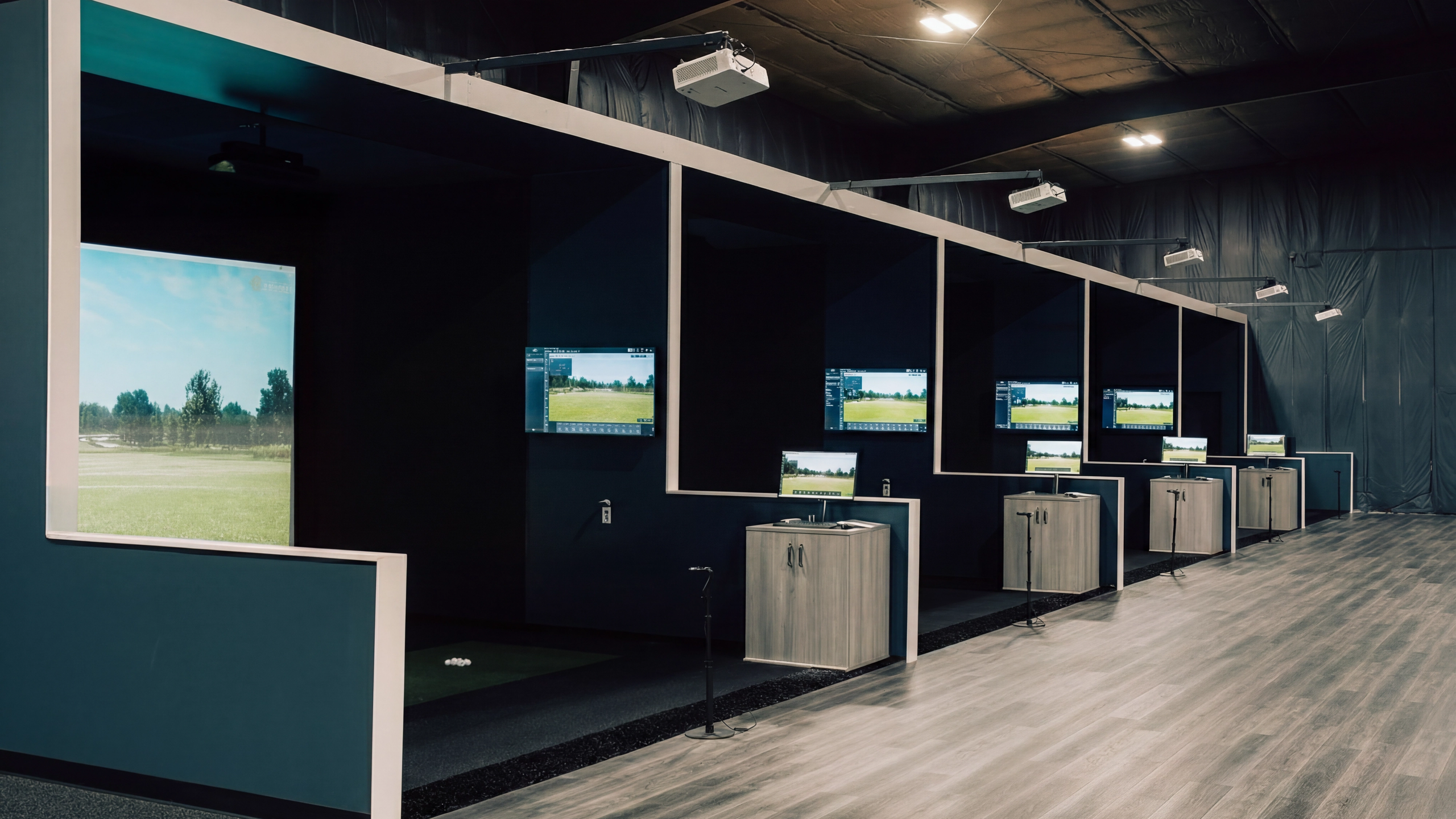 A row of indoor golf simulator bays, each with a large screen showing a golf course, a hitting mat, and a data monitor.