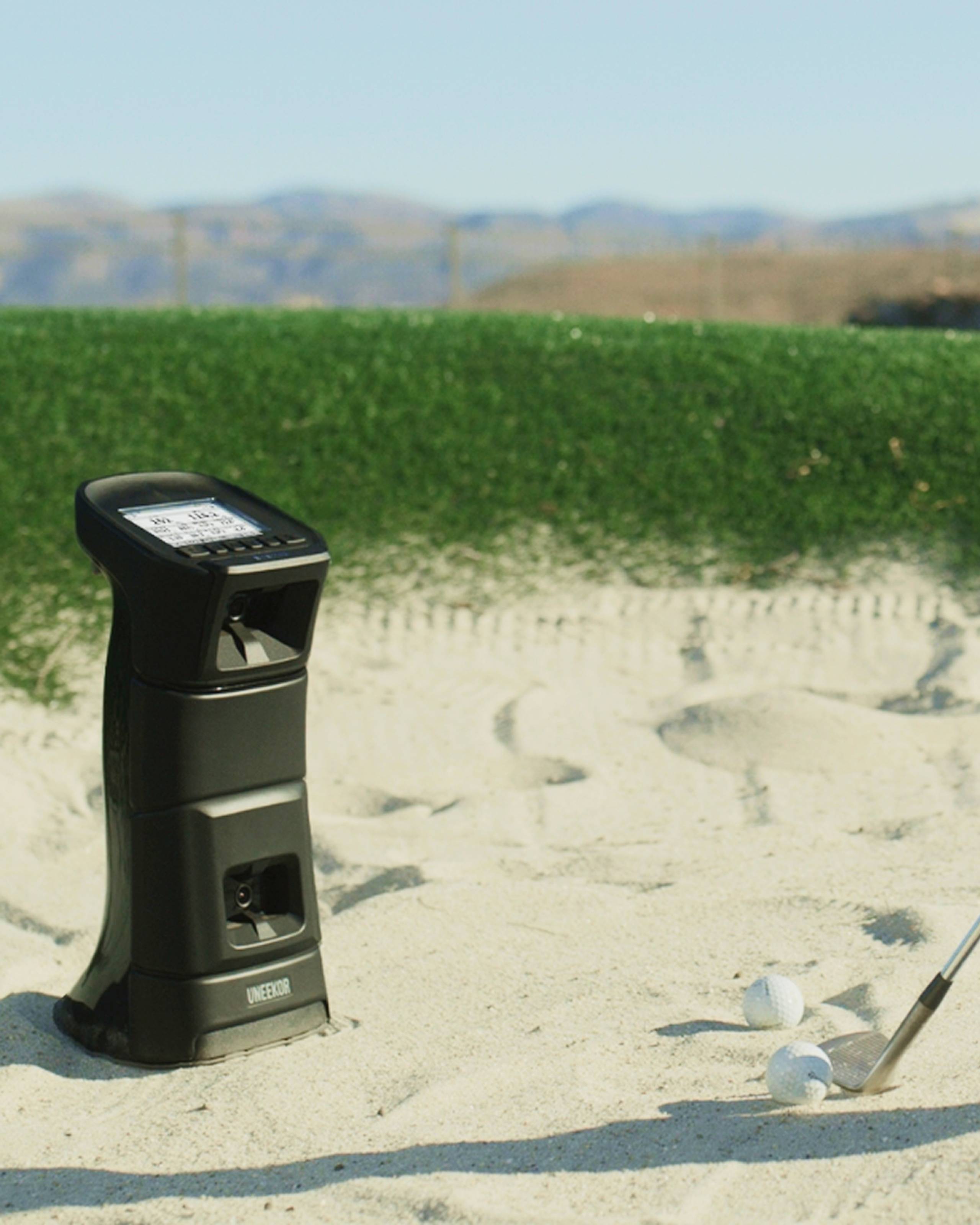A black golf launch monitor in a sand trap with two golf balls and a golf club.