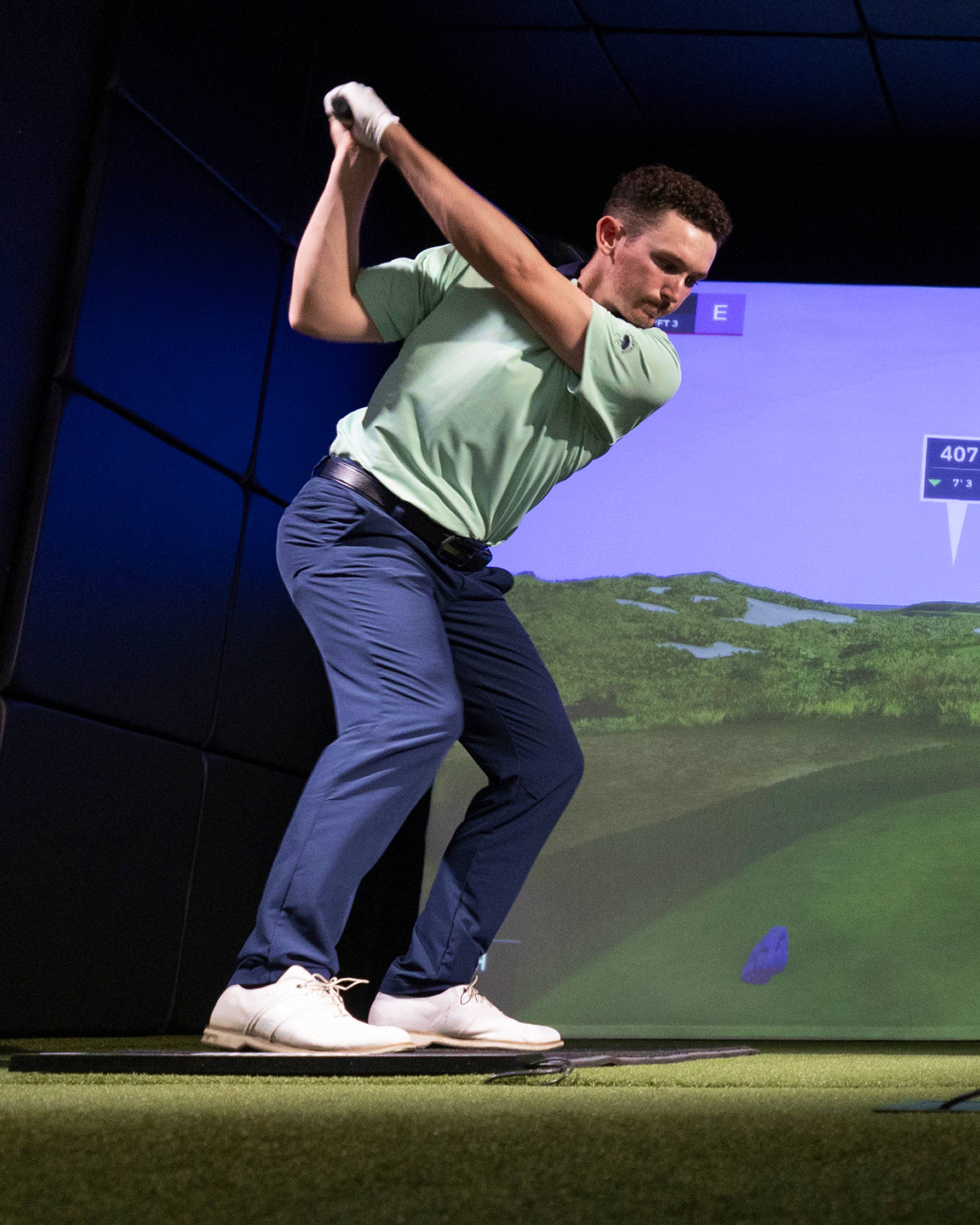Man in green polo and blue pants mid-golf swing in an indoor simulator, with a virtual golf course on screen.
