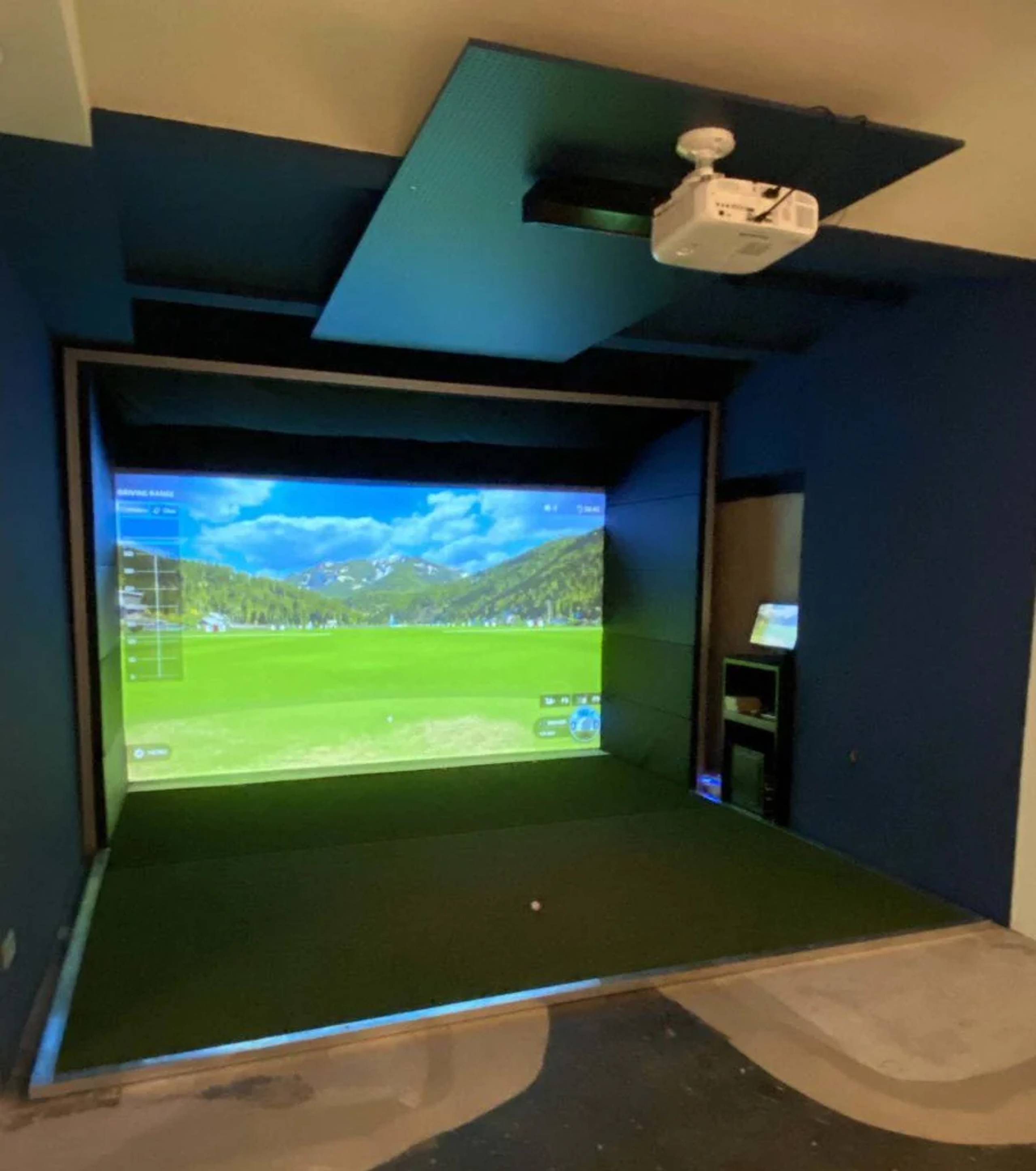 Golf simulator with a virtual course projected onto a large screen, a projector, and a golf ball on a green hitting mat.