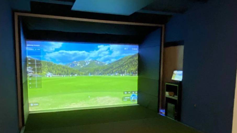 Golf simulator with a virtual course projected onto a large screen, a projector, and a golf ball on a green hitting mat.
