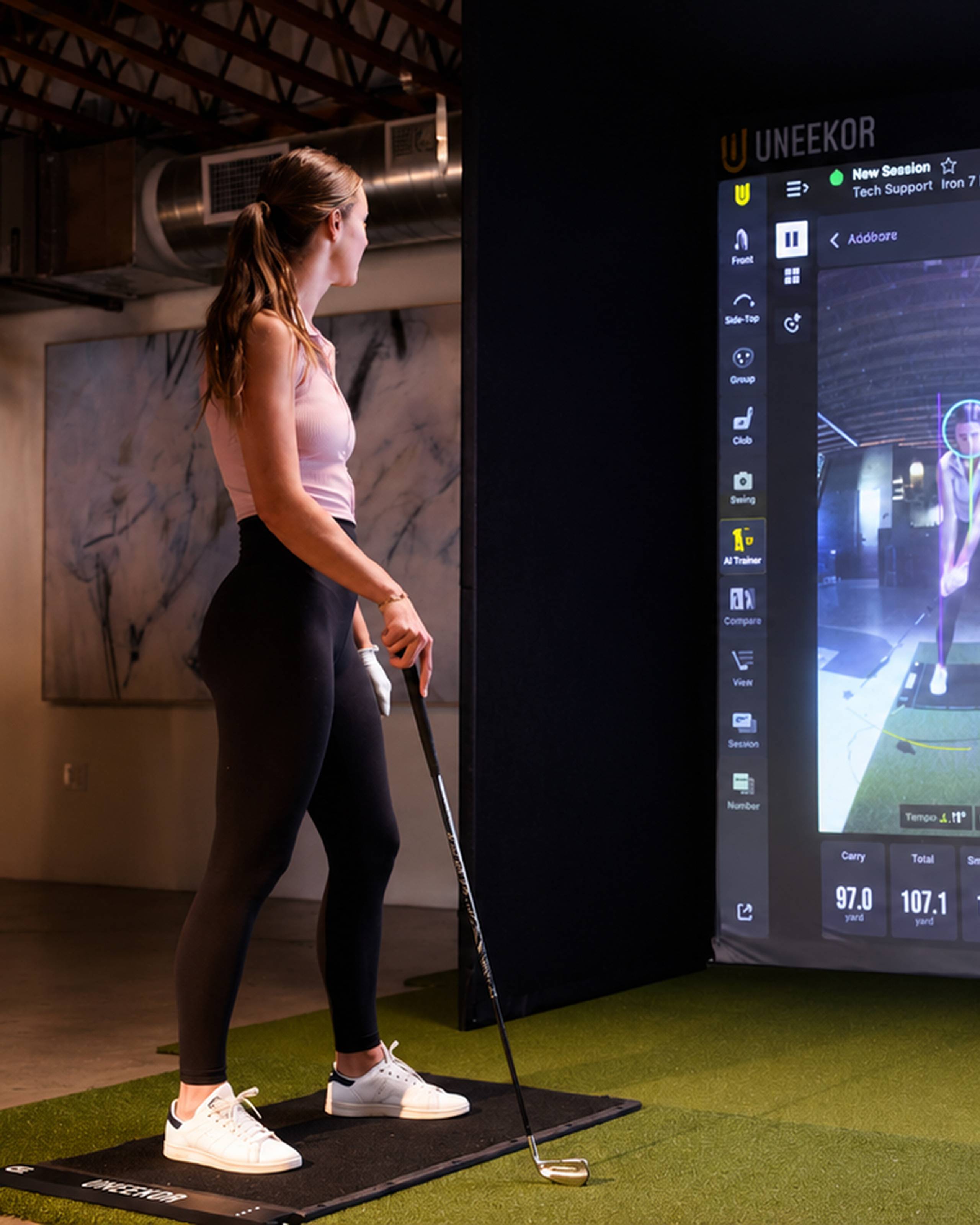 A woman in athletic wear holds a golf club while looking at a UNEEKOR golf simulator screen.