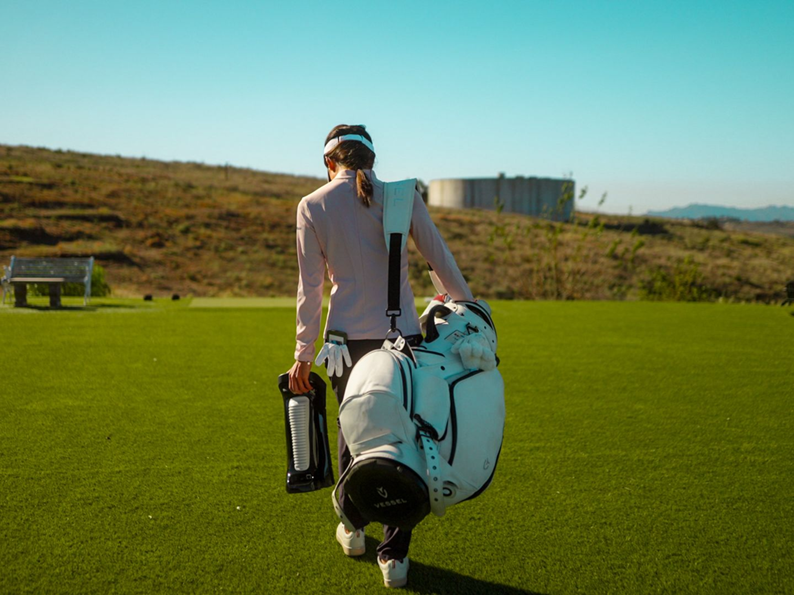 A golfer walks away on a green, carrying a white golf bag and a black insulated bottle.