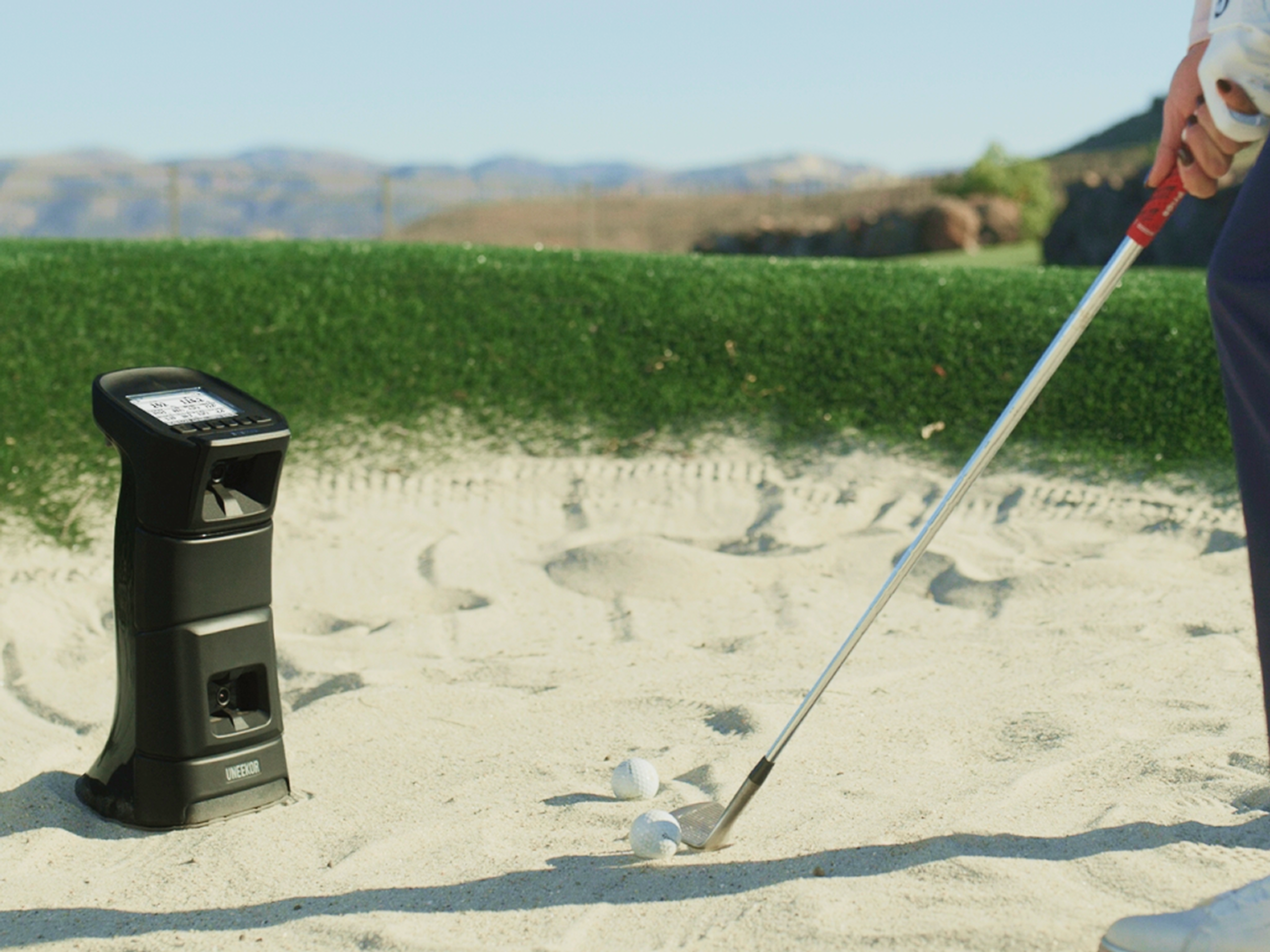 A golfer prepares to hit a golf ball from a sand bunker, with a black golf launch monitor set up nearby.