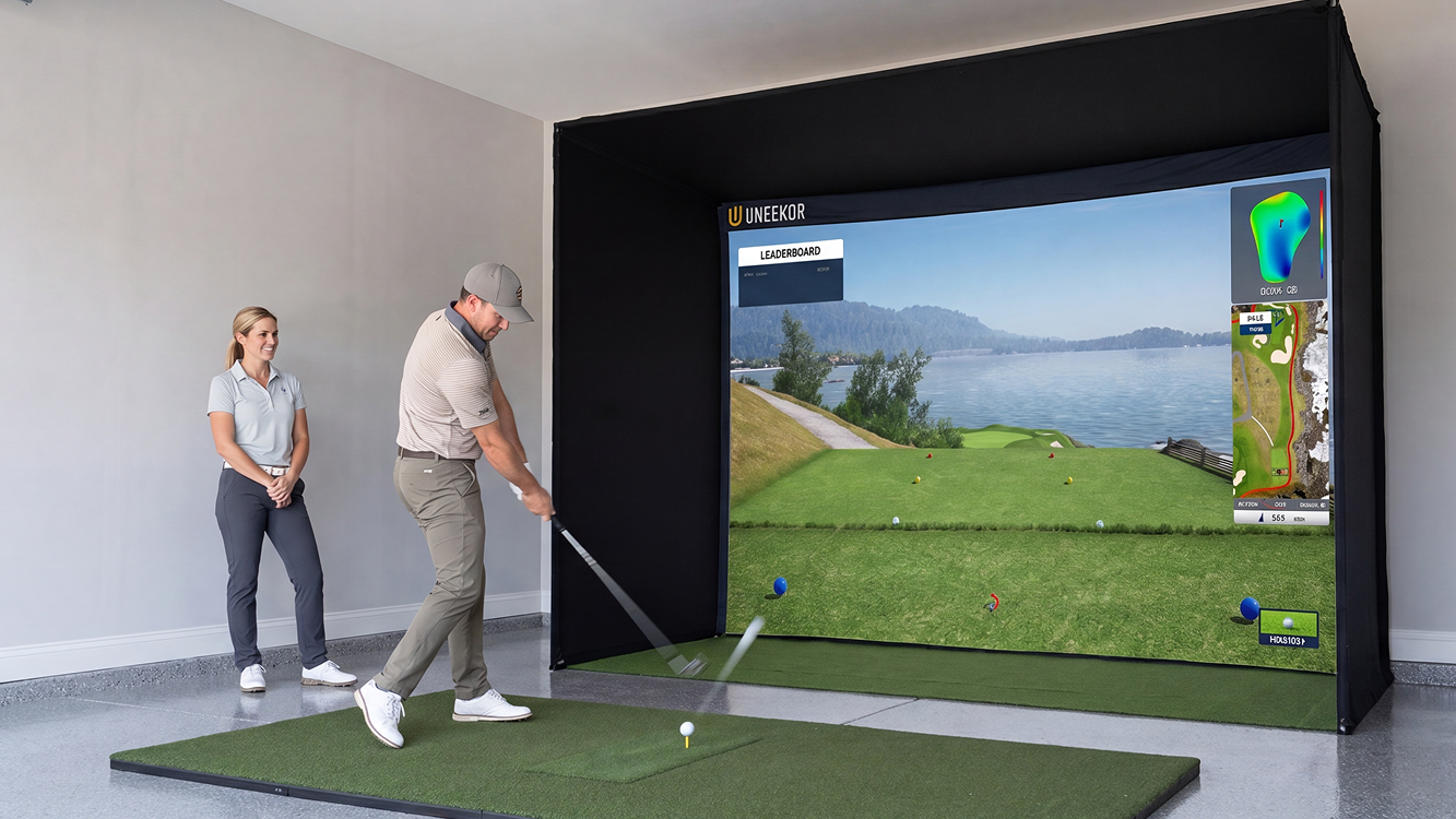 A man swings a golf club on an indoor golf simulator in a garage, with a woman watching.