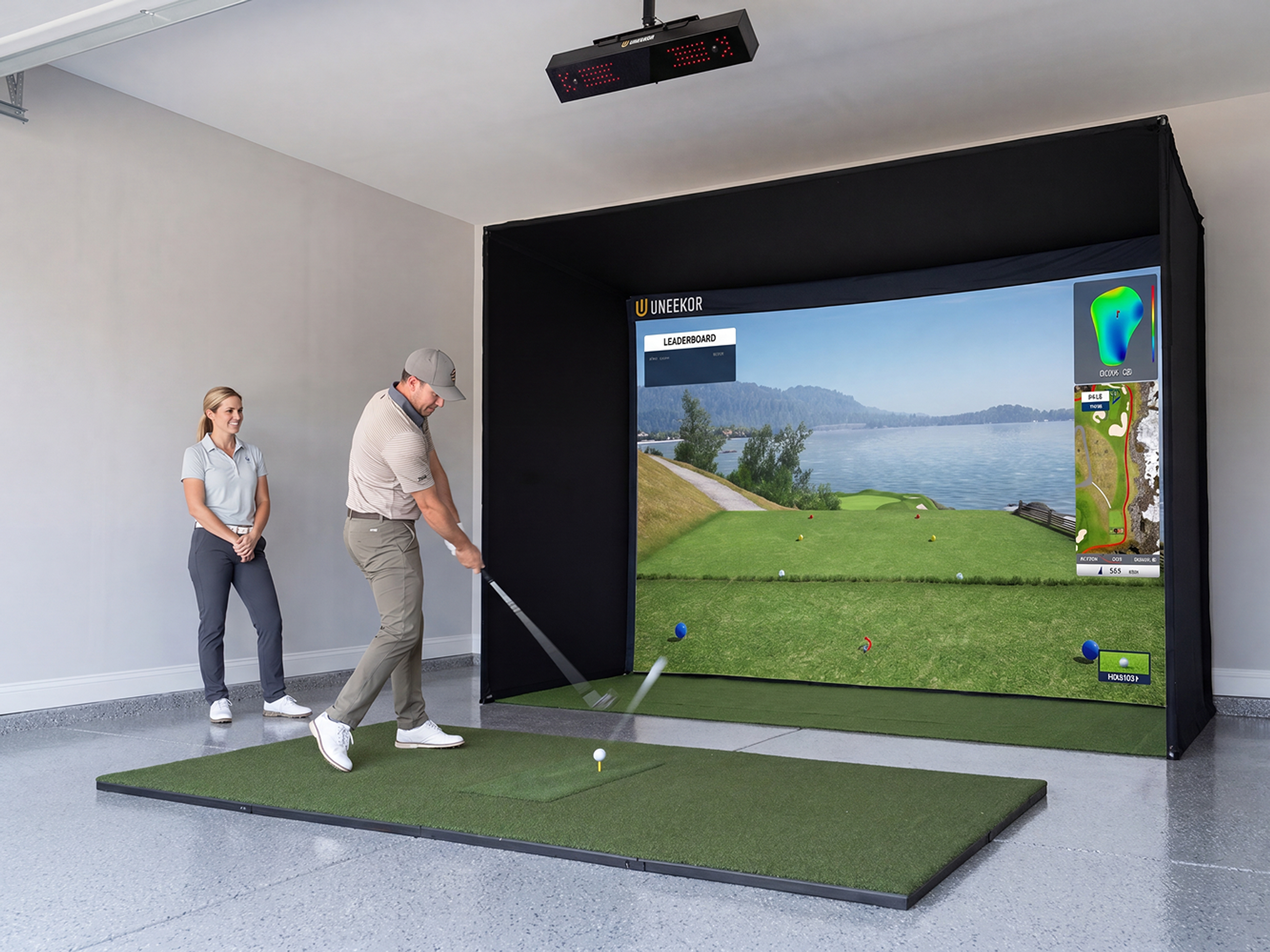 A man swings a golf club on a golf simulator setup in a garage, watched by a woman.