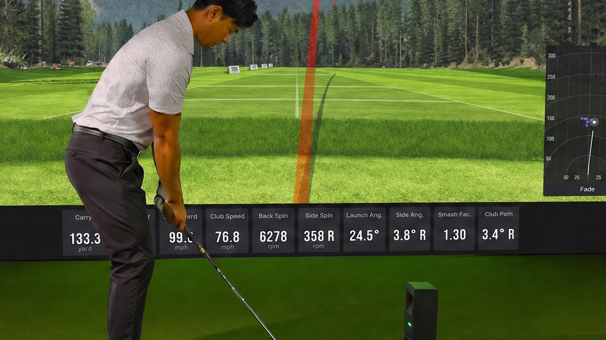 A golfer at a simulator with a virtual mountain course, red ball trajectory, and swing stats on screen.