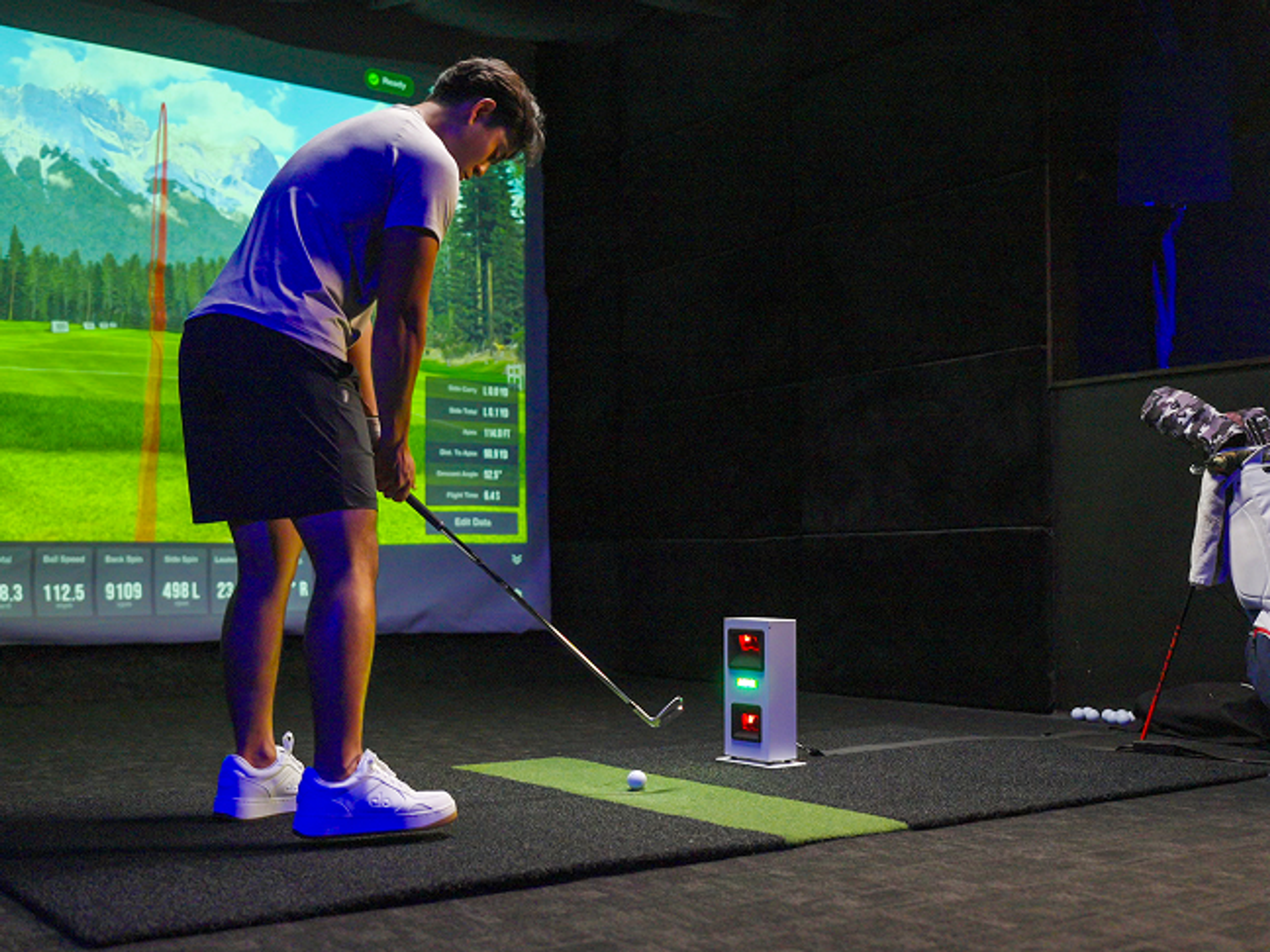 A man plays golf in an indoor simulator, aiming at a virtual course on a screen with a launch monitor.