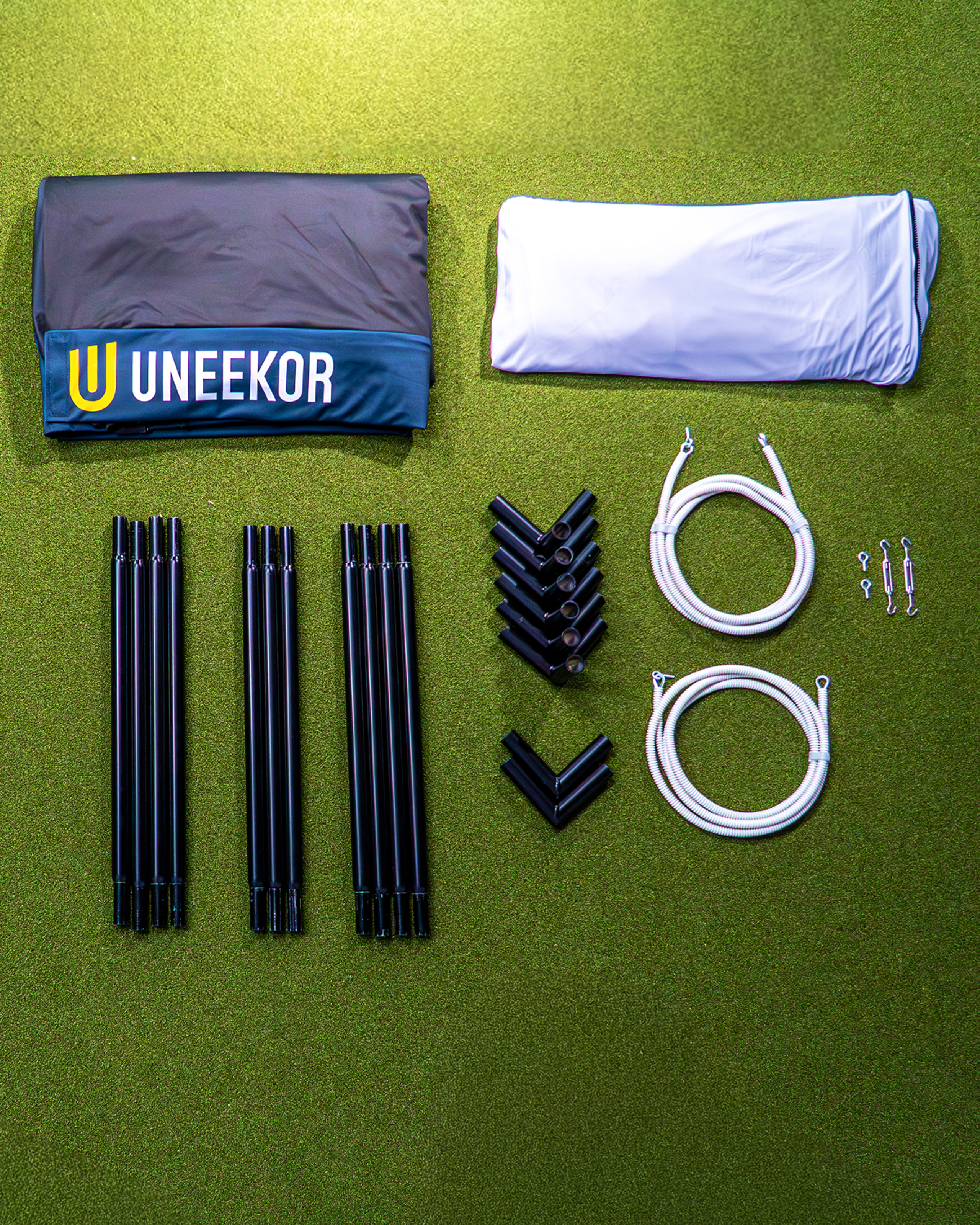 Unassembled Uneekor golf simulator kit components, including a black enclosure, white screen, black frame poles, connectors, and ropes, laid out on a green surface.