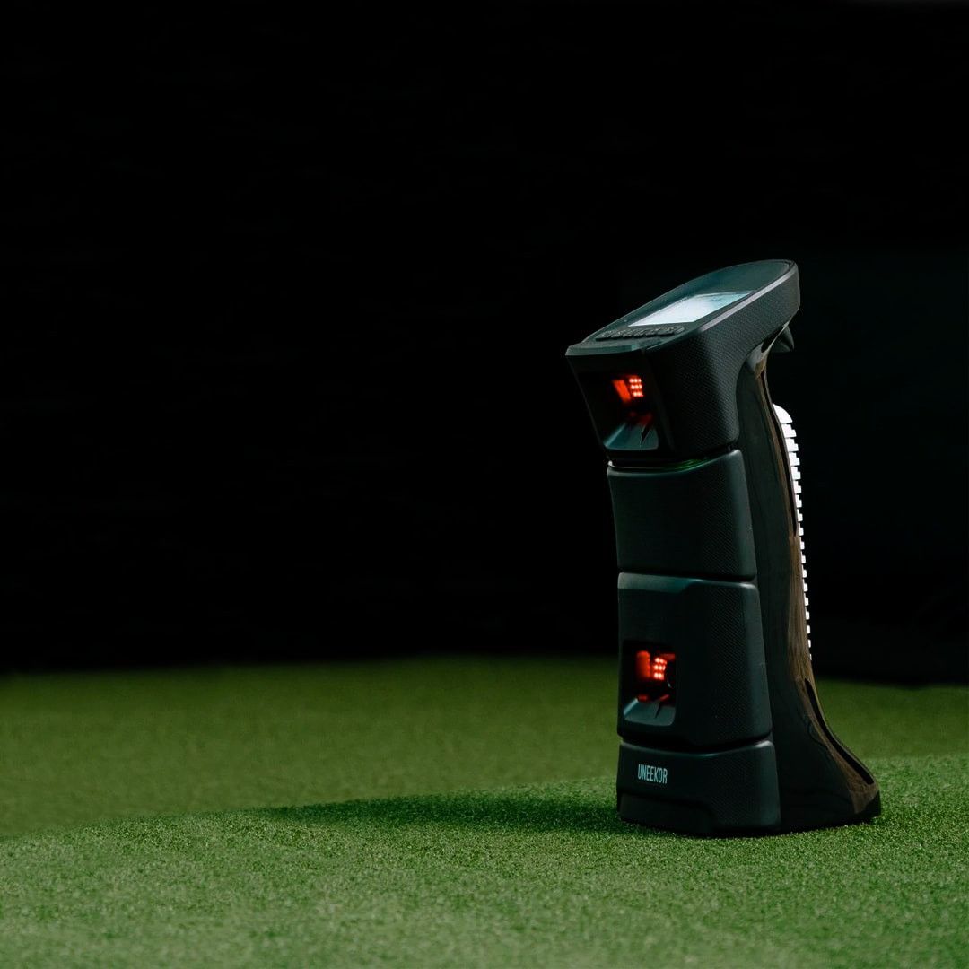 A dark UNIEQR golf launch monitor with red lights and a screen sits on green turf.
