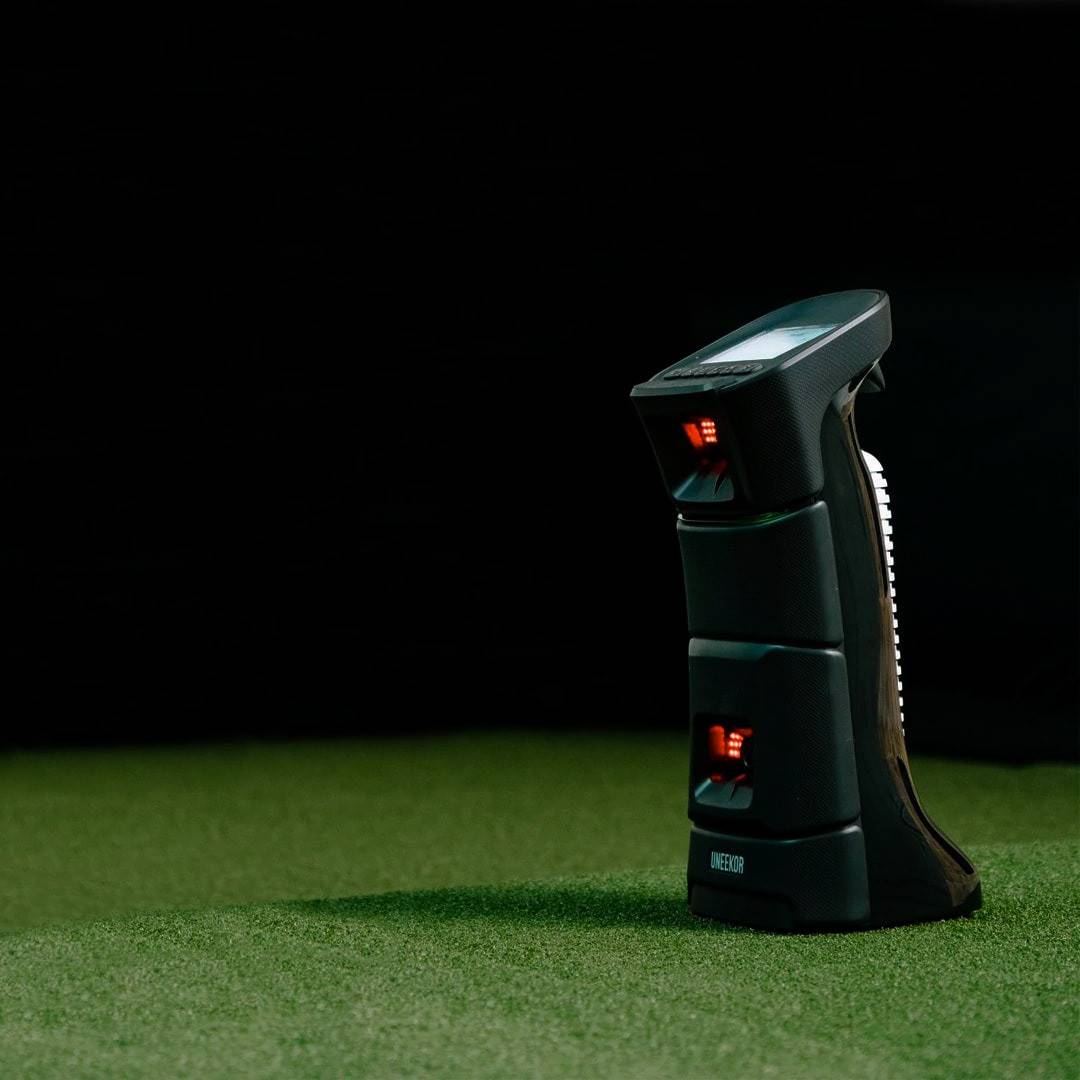 A dark UNEKOR golf launch monitor with glowing red sensors sits on green artificial grass.