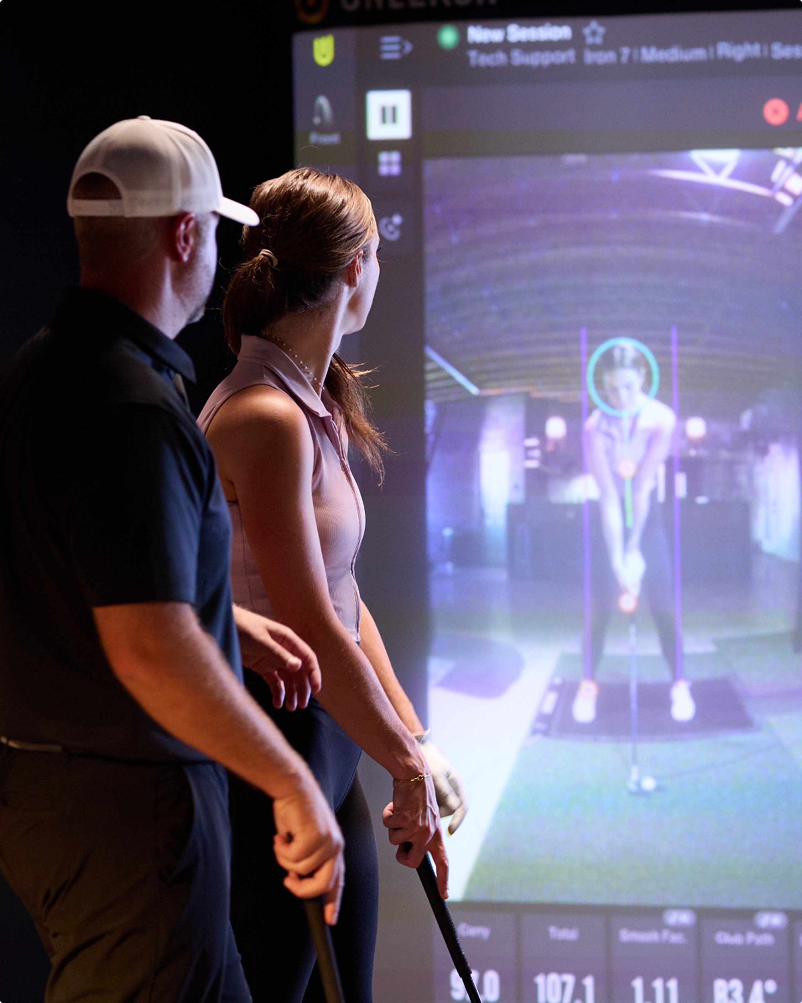 Two golfers observe a large screen displaying a golf simulation with tracking data.