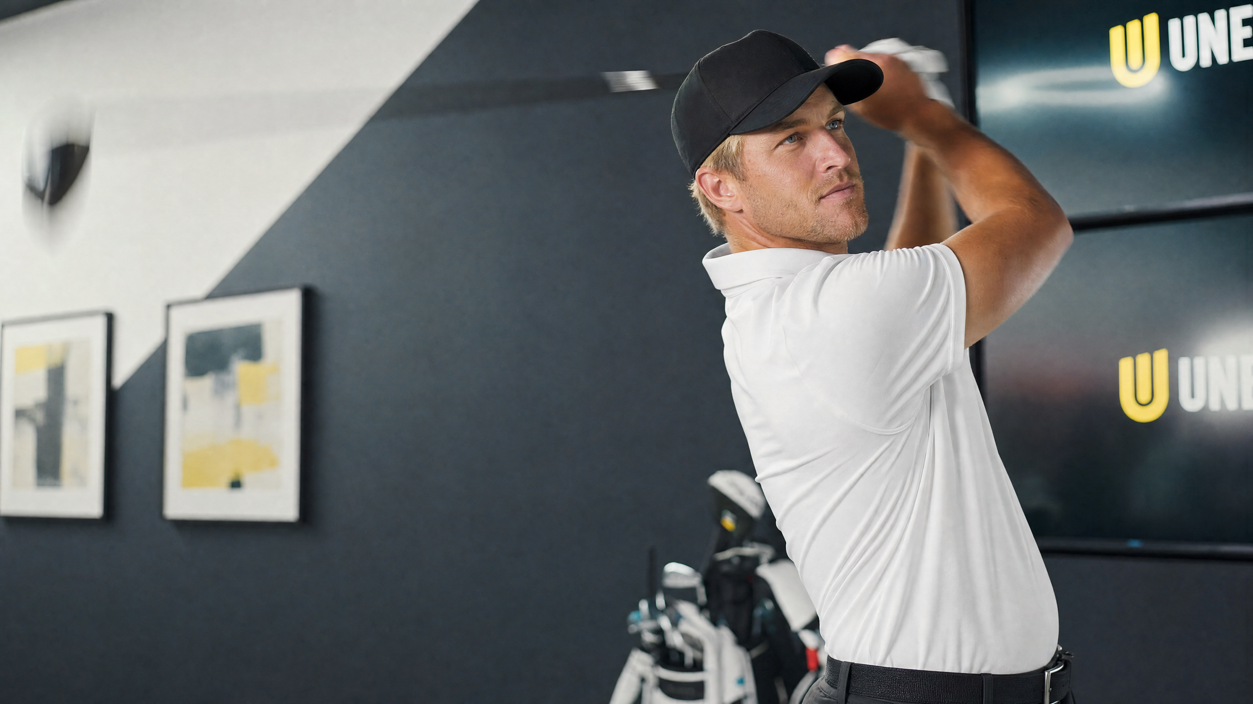 A man in a black cap and white polo shirt mid-swing with a golf club indoors.