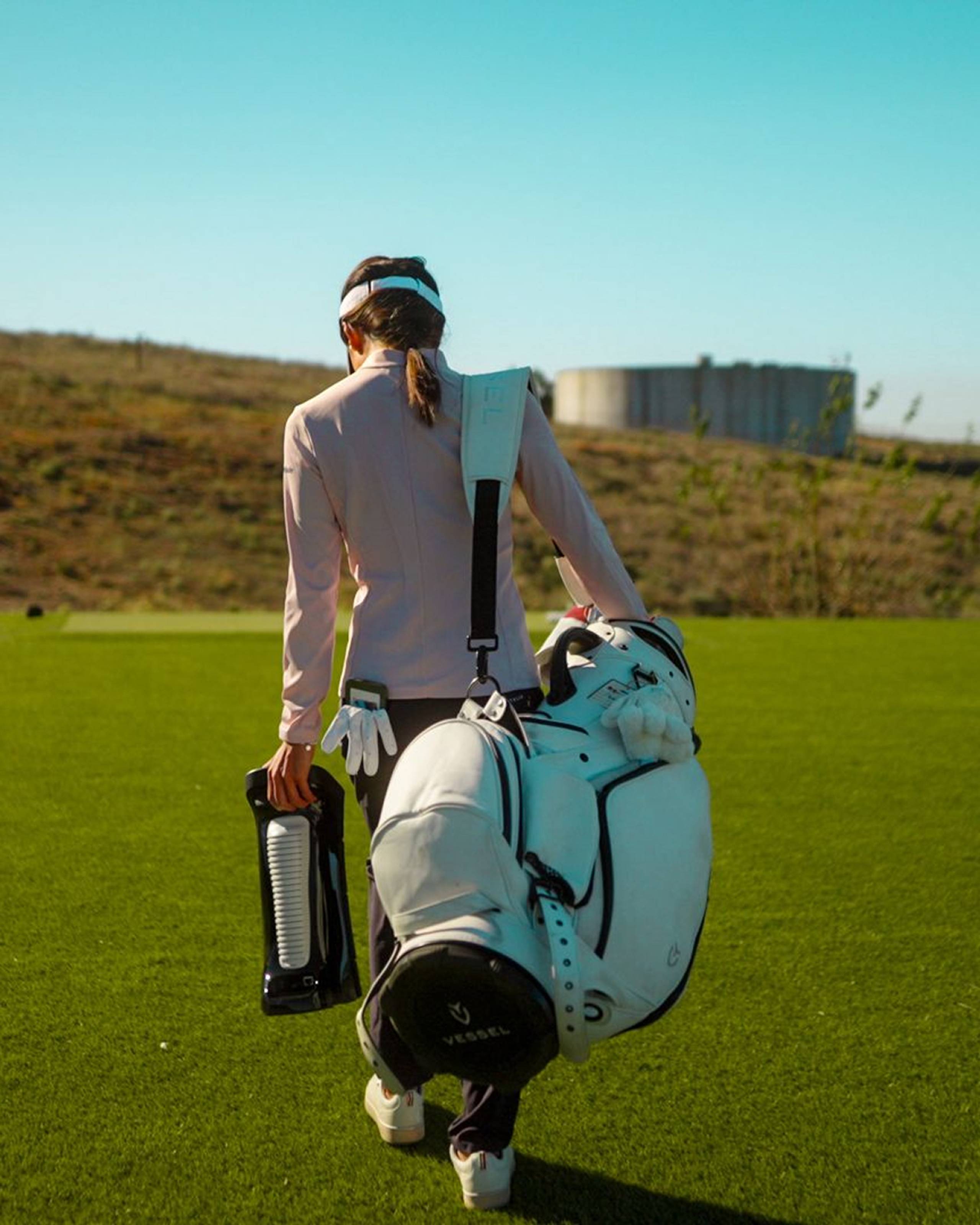 A golfer walks away across a green course, carrying a white golf bag and a black hydration device.