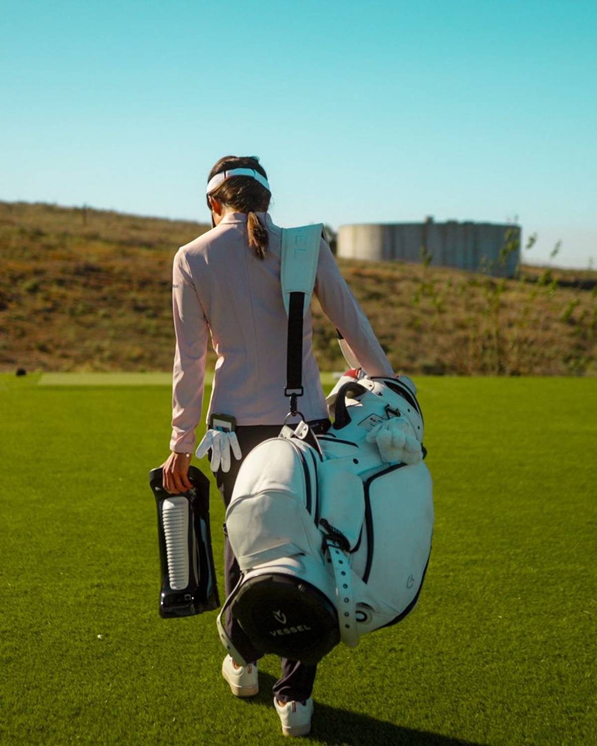 A golfer walks away across a green course, carrying a white golf bag and a black hydration device.