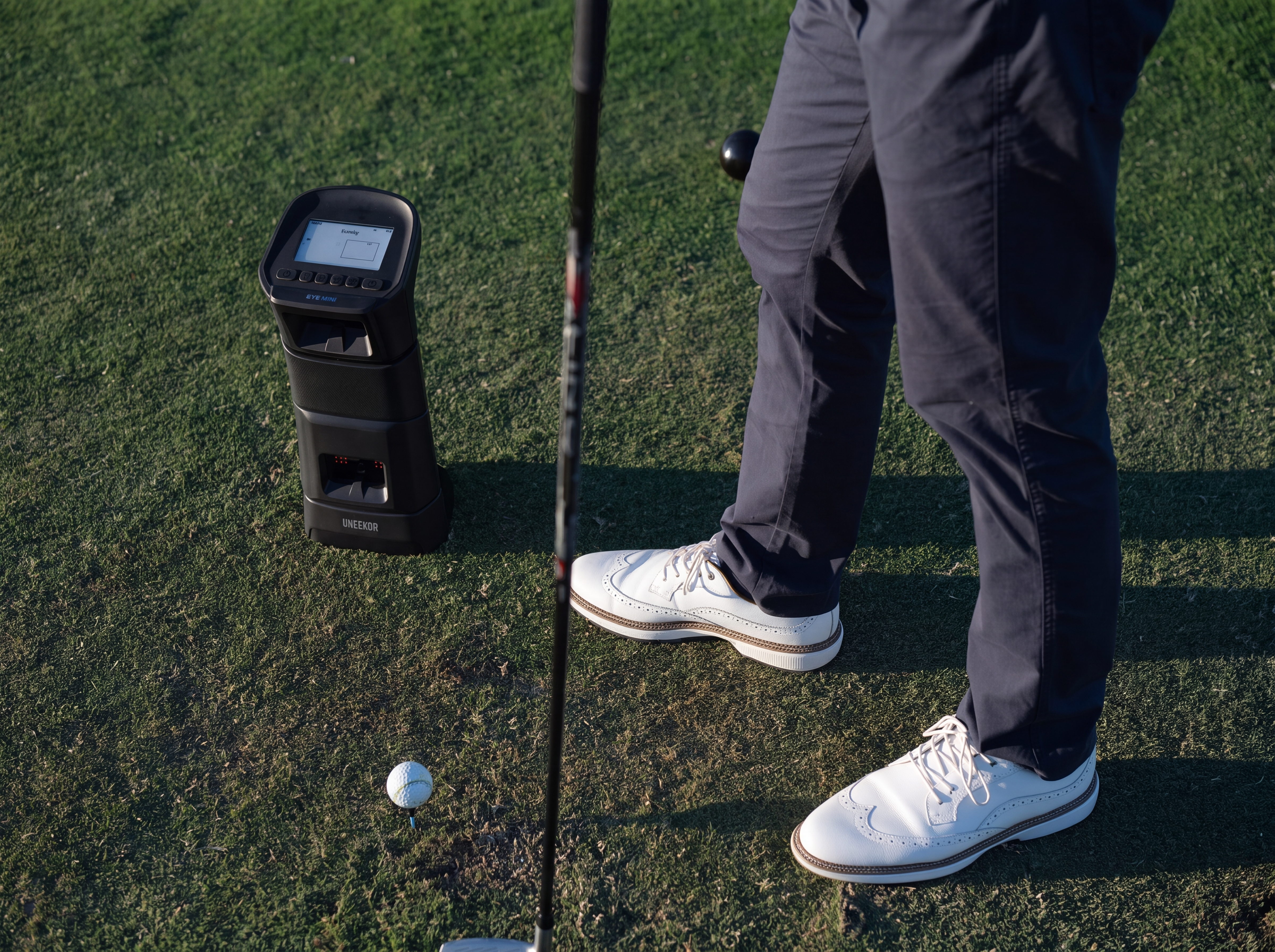 A golfer's legs and feet stand on a green next to a golf ball, club, and a black golf launch monitor.