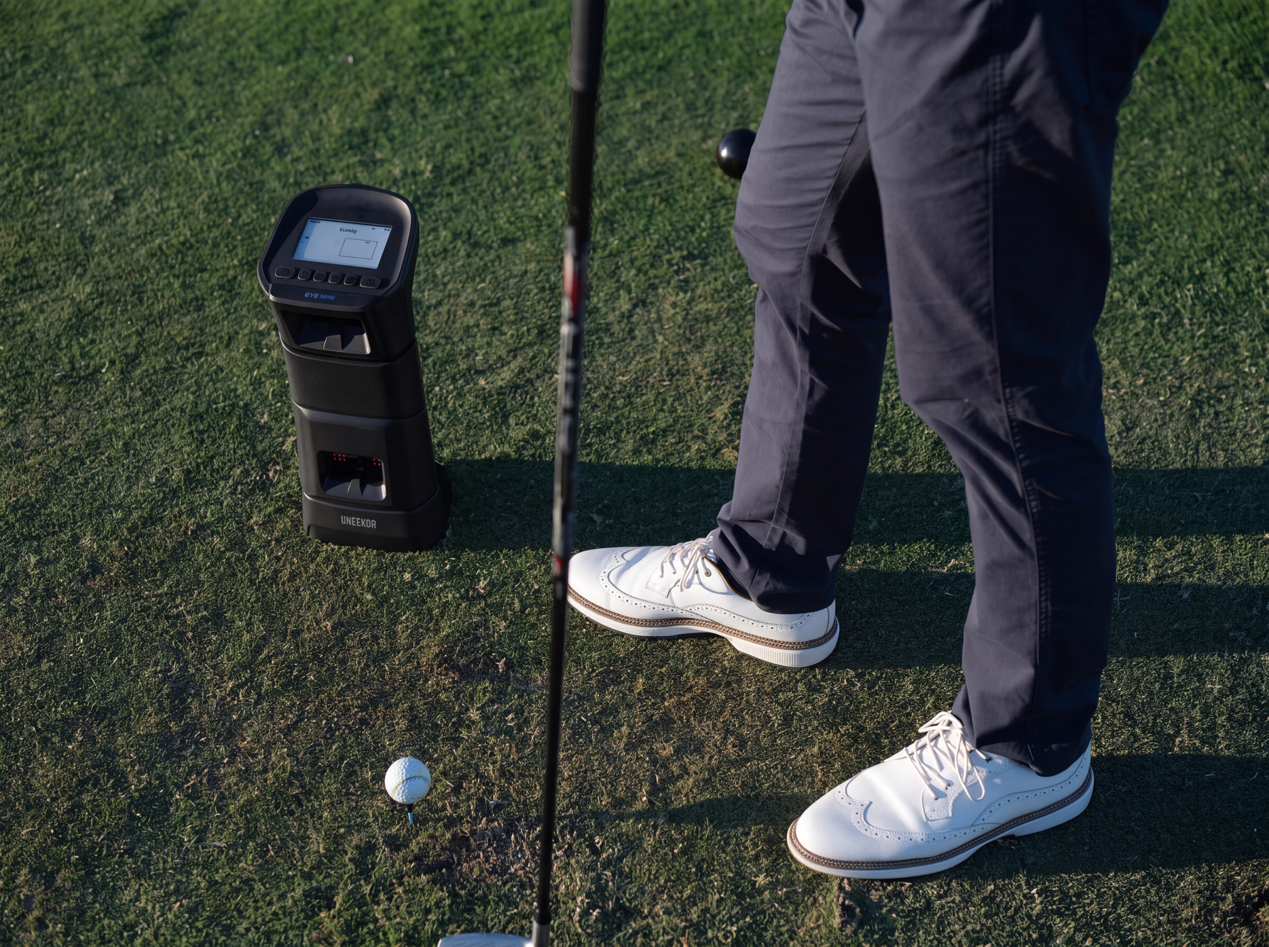A golfer's legs and feet stand on a green next to a golf ball, club, and a black golf launch monitor.