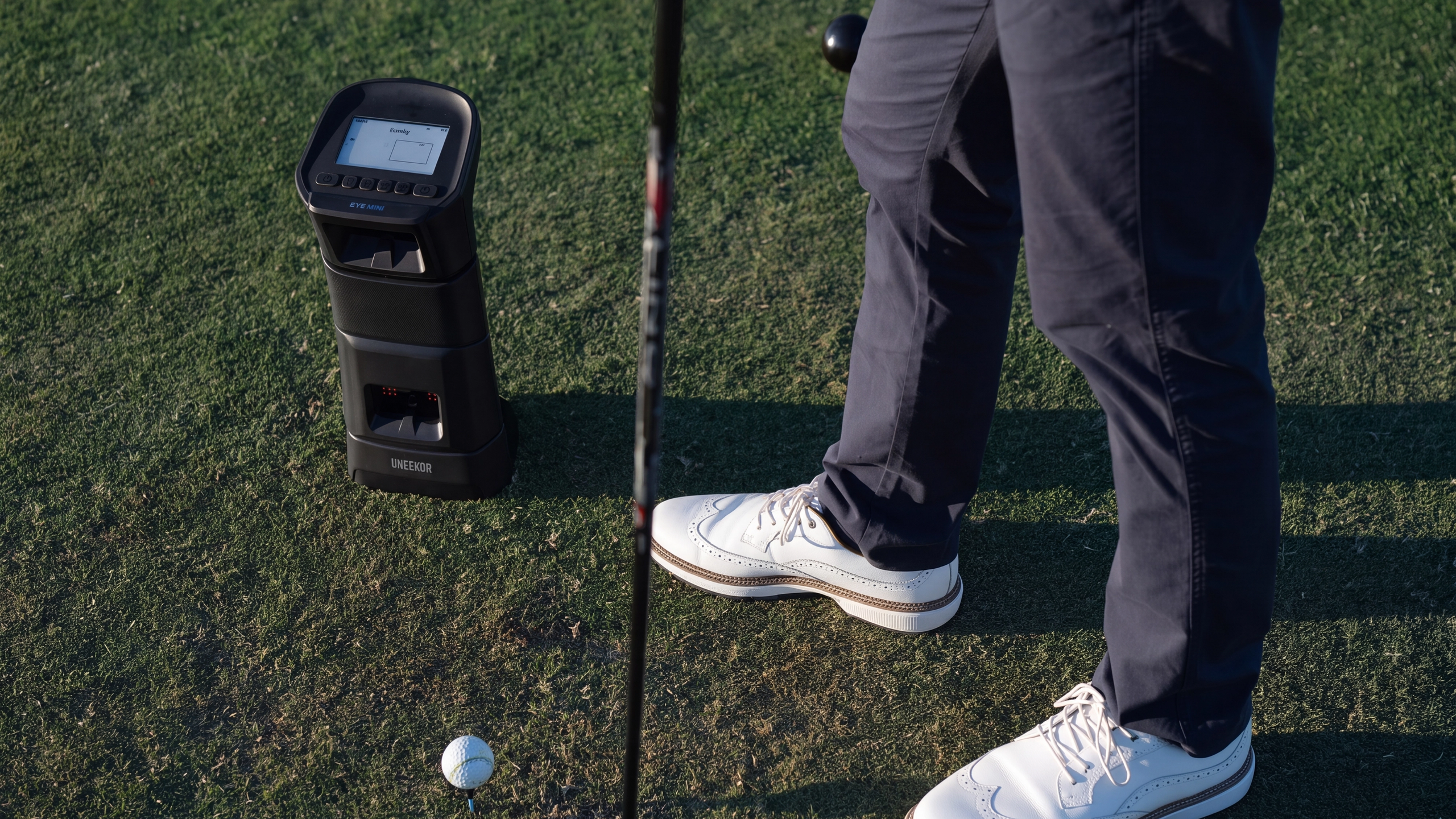 A golfer's legs and feet stand on a green next to a golf ball, club, and a black golf launch monitor.
