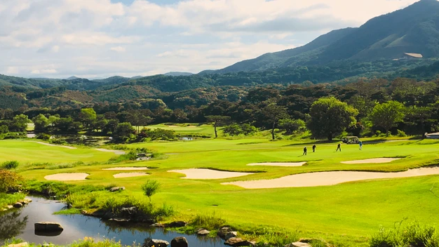 A green golf course with sand traps, a stream, golfers, and forested mountains under a cloudy sky.
