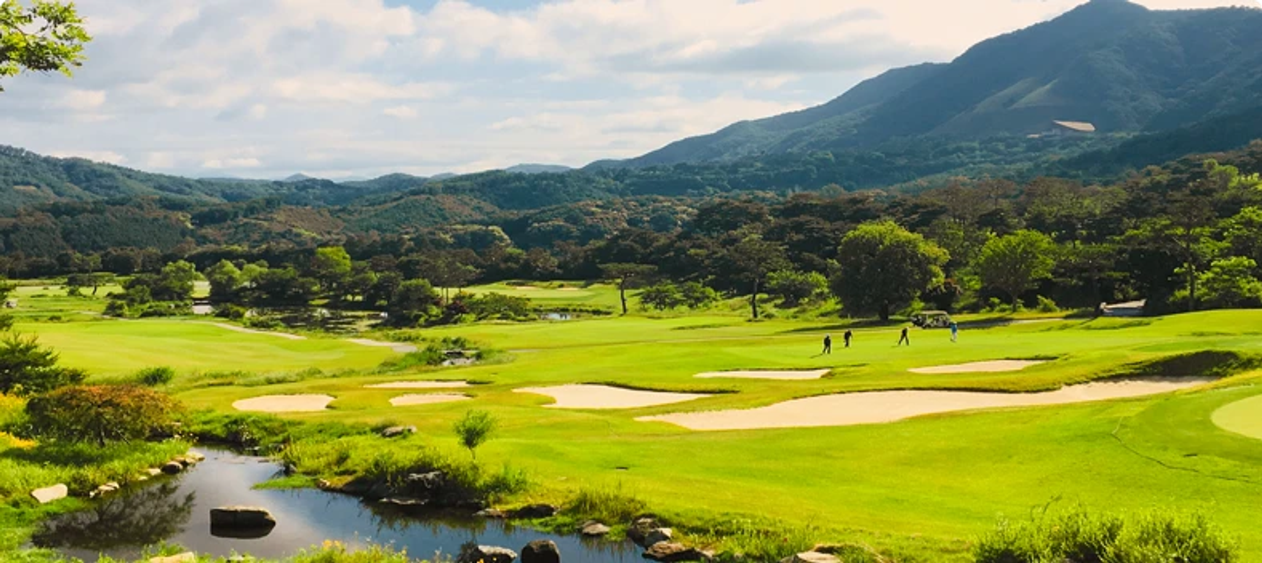 A wide view of a golf course with green fairways, sand traps, a stream, golfers, and distant mountains.