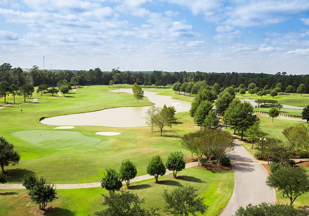 Aerial view of a green golf course with winding water features, abundant trees, and cart paths under a partly cloudy sky.