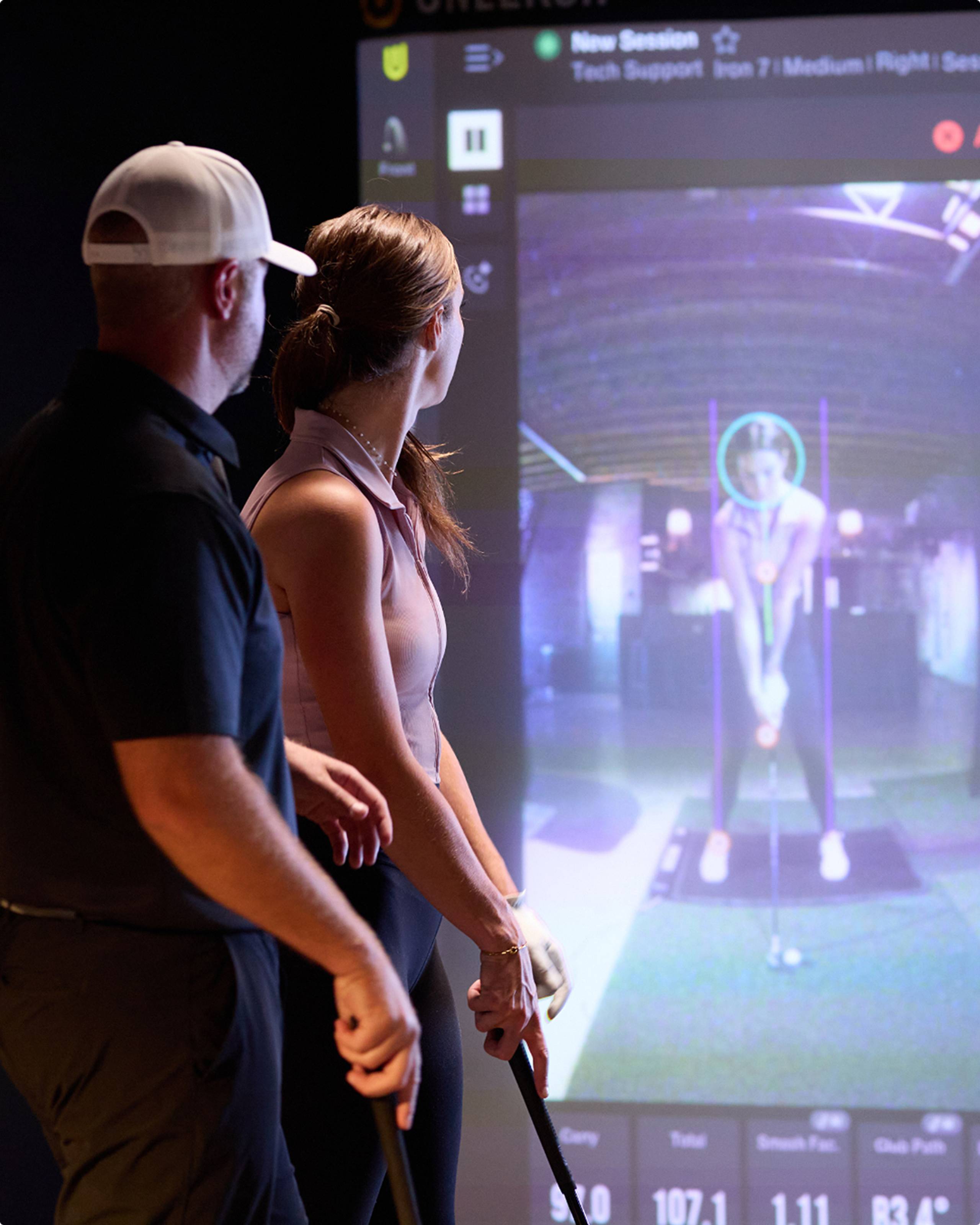 A man and a woman watch a golf simulator screen displaying a golfer and swing analysis, both holding golf clubs.
