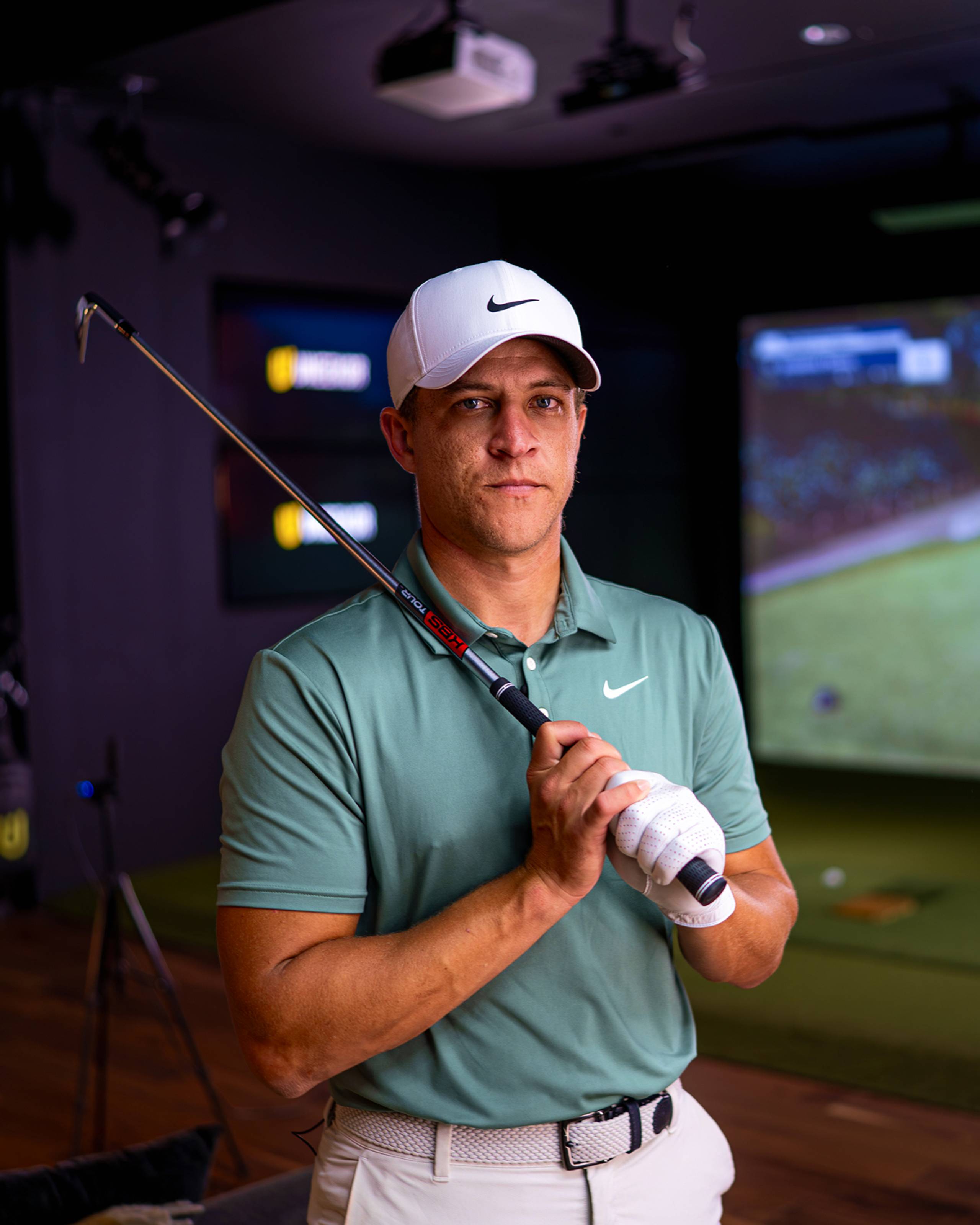 A man in a white Nike cap, green Nike polo, and white golf glove holds a golf club in an indoor golf simulator.