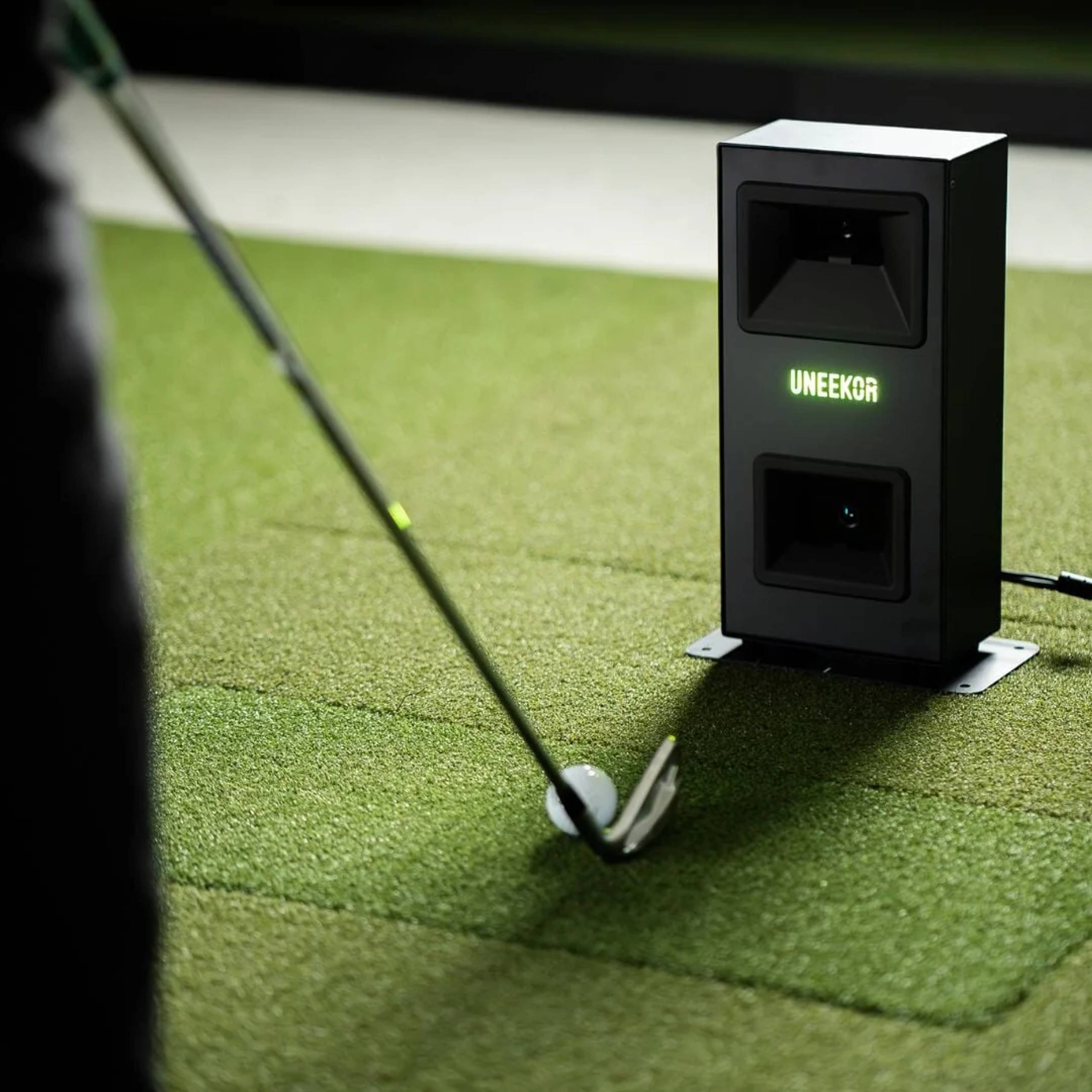 A golf club addresses a golf ball on green turf, next to a black Uneekor golf launch monitor.