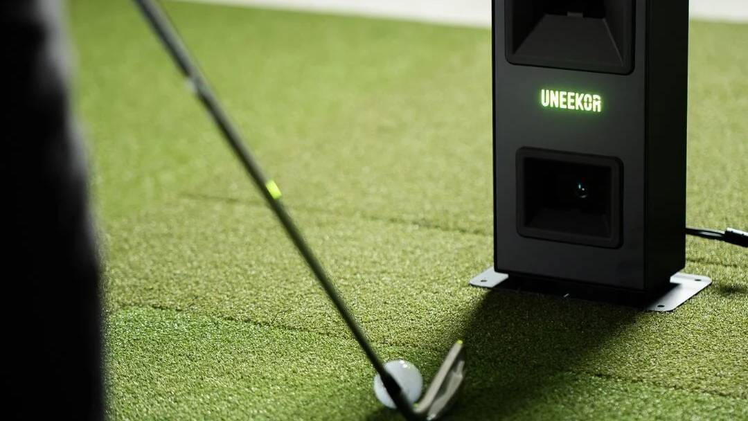 A golf club addresses a golf ball on green turf, next to a black Uneekor golf launch monitor.