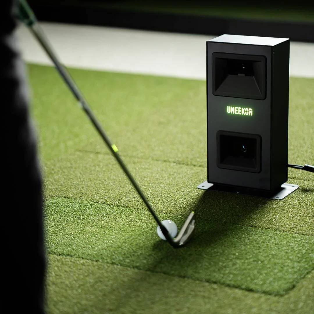 A golf club addresses a golf ball on green turf, next to a black Uneekor golf launch monitor.