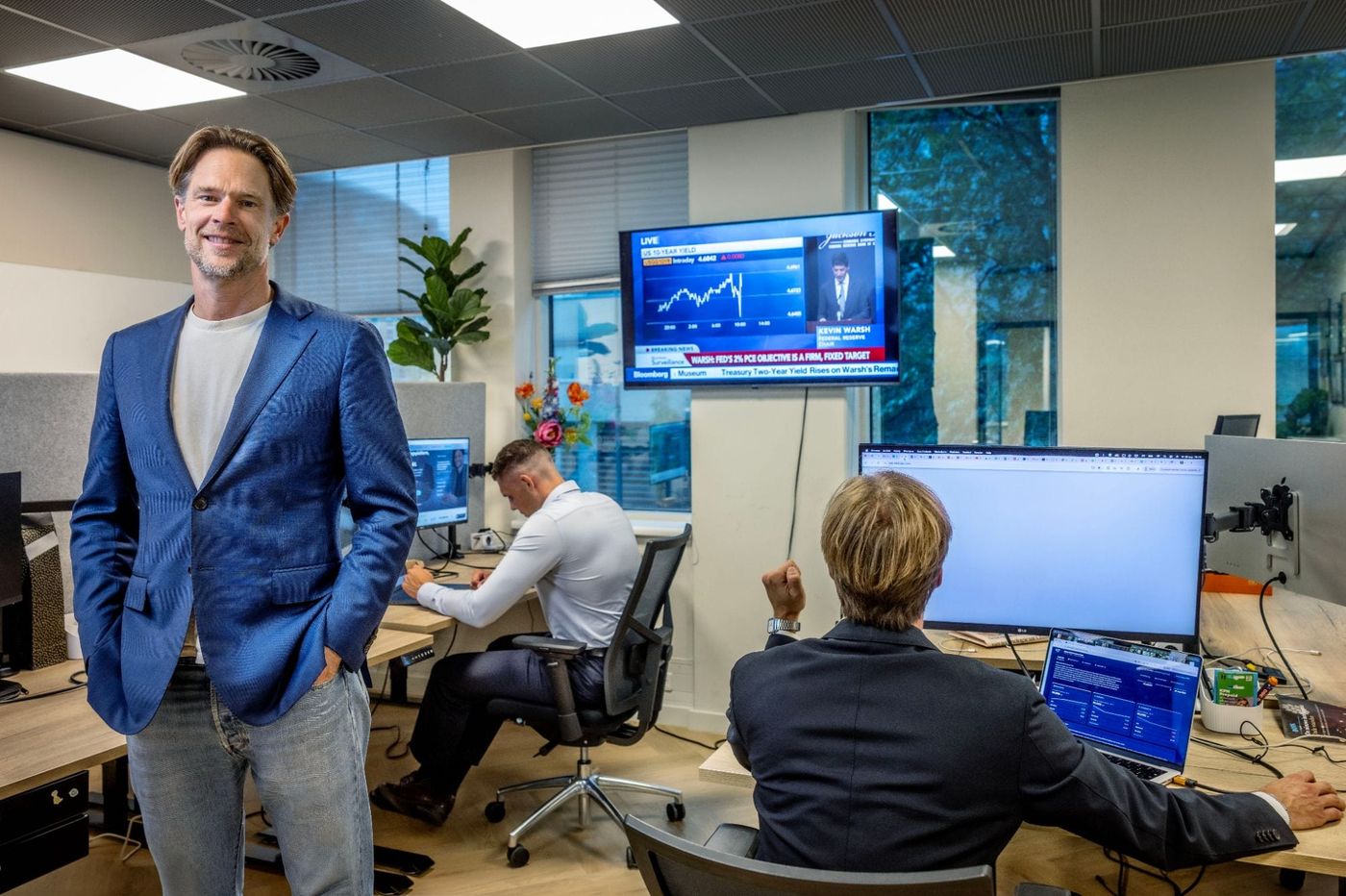 Michiel Stokman op het kantoor van BOTS Capital, met achter hem het team aan het werk en een beursscherm aan de wand