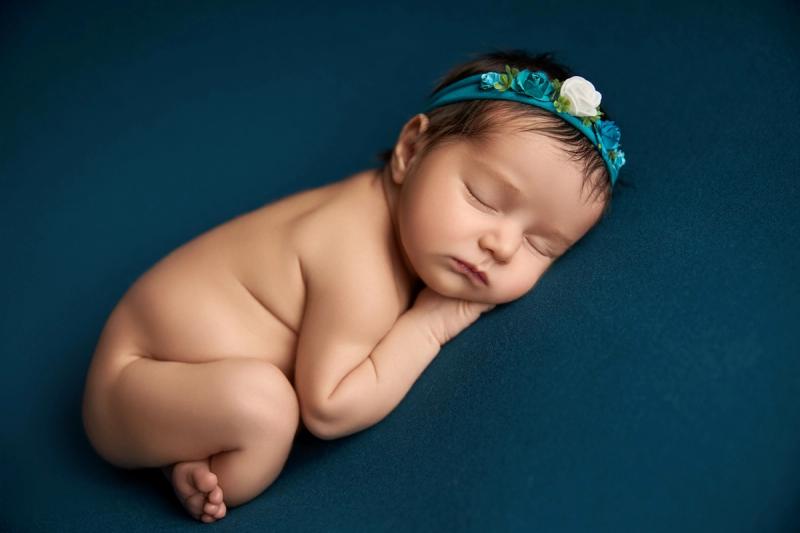 Novorozená holčička na tmavě modré dece s tyrkysovou čelenkou.
