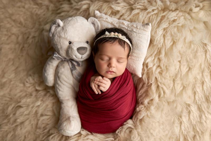 Newborn holčička v červeném wrapu s medvídkem na polštářku.