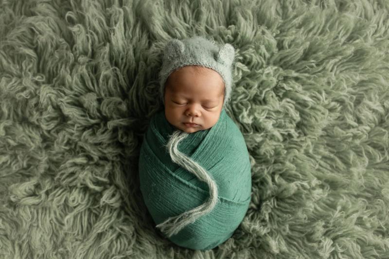 Miminko v zeleném wrapu s medvědí čepičkou na zelené kožešině.