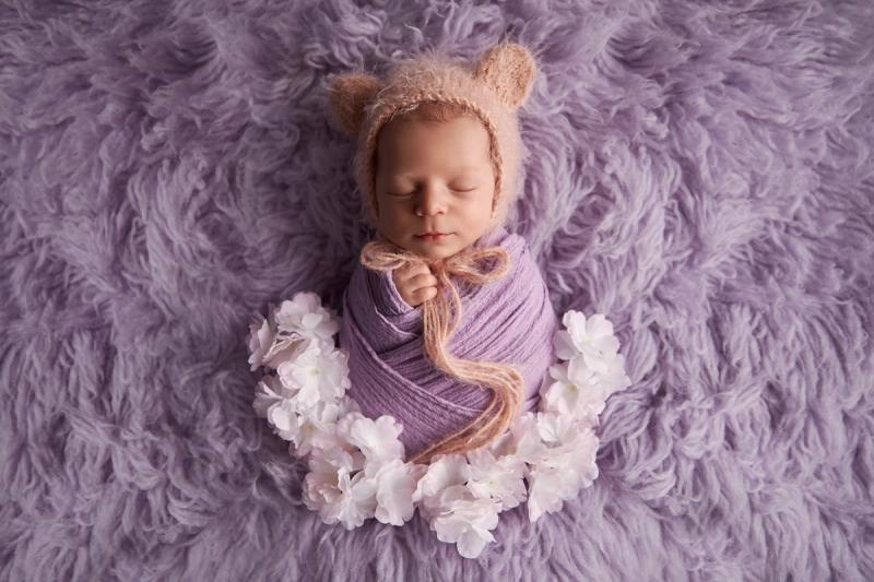 Novorozená holčička ve fialovém wrapu s medvědí čepičkou a věncem.