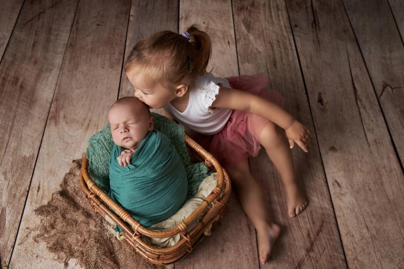 Holčička dává pusu novorozenci v košíčku na dřevěné podlaze.