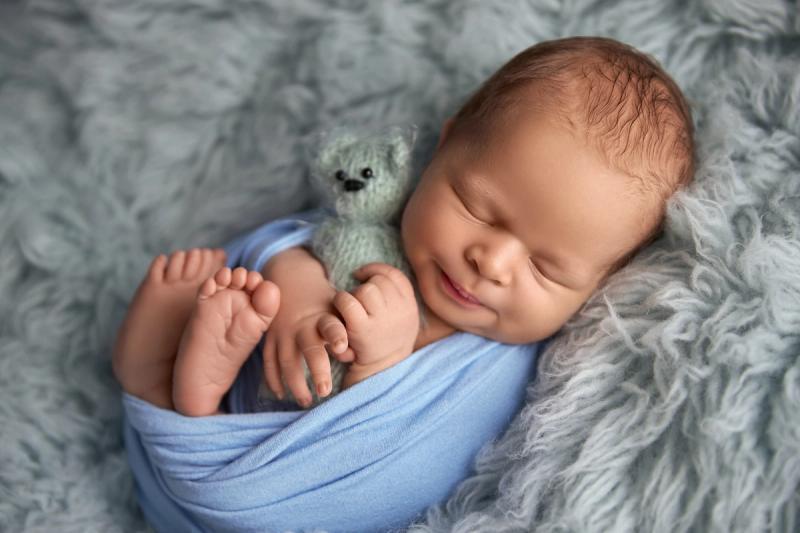 Newborn miminko v modrém wrapu s úsměvem a medvídkem.
