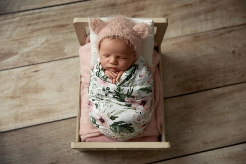 Novorozená holčička v dřevěné postýlce s růžovou čepičkou.