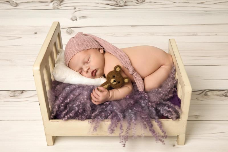Newborn holčička v dřevěné postýlce se starorůžovou čepičkou a medvídkem.