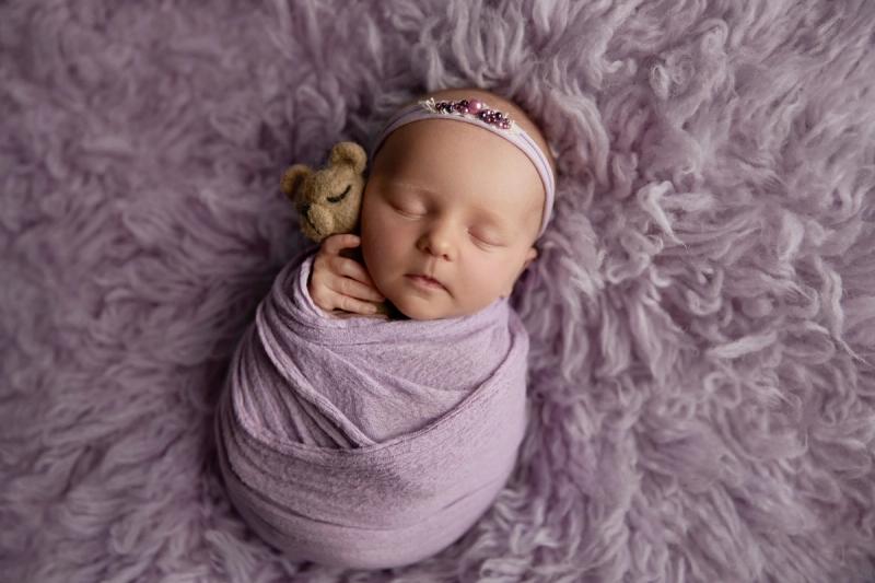 Newborn holčička ve fialovém wrapu s medvídkem a čelenkou.