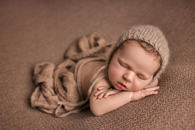 Miminko v béžovém wrapu a čepičce na bříšku na hnědém pozadí.