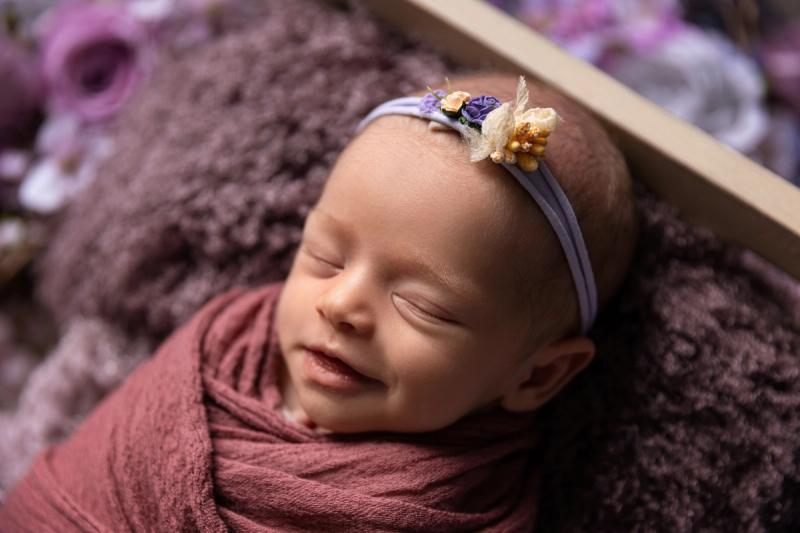 Newborn holčička s úsměvem ve starorůžovém wrapu v dřevěné postýlce.