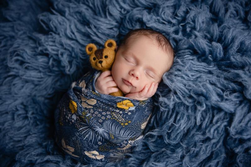 Novorozenec v tmavě modrém wrapu s medvídkem na modré kožešině.
