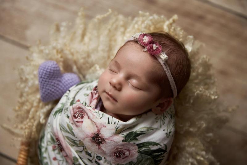 Novorozená holčička v košíku s květinovým wrapem a fialovým srdcem.