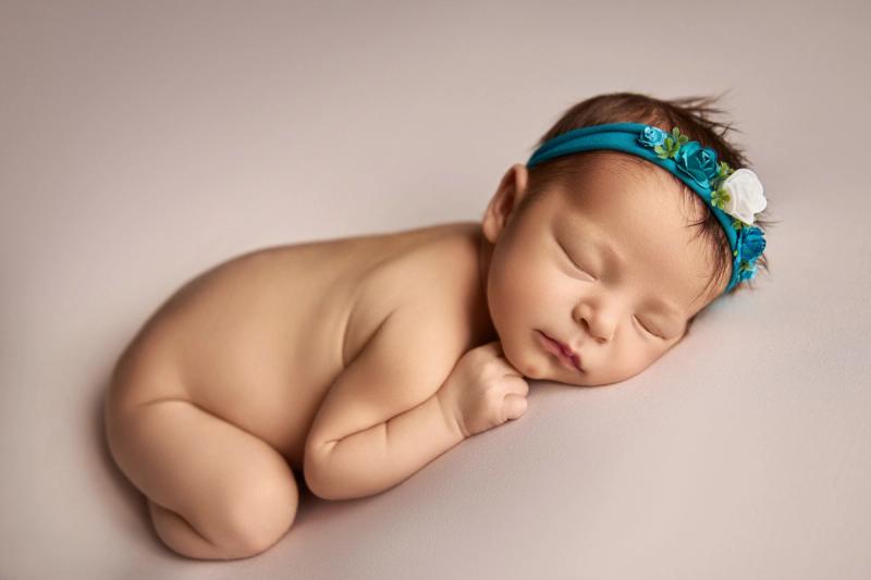 Novorozená holčička na bříšku bez oblečení s tyrkysovou čelenkou.